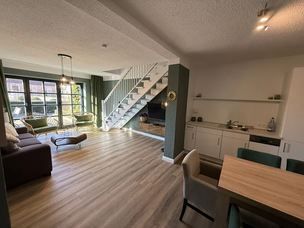 Lichte woonkamer met open keuken, trap naar de bovenverdieping en grote ramen met uitzicht op de tuin.
