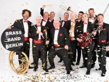 Elf Musiker der Brass Band Berlin posieren fröhlich mit Metallblasinstrumenten und Konfetti.