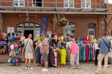 Menschen stöbern vor einem Backsteingebäude durch bunte Kleidung in einem Straßenverkauf.