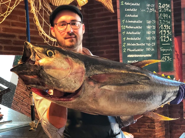 Frischfisch bei Fiedler IMG_2118 (c) Fiedler_Erlebnis Bremerhaven.jpeg Mann mit Brille und Mütze hält großen Thunfisch in Fischmarkt mit Schiefertafel und Preisen.