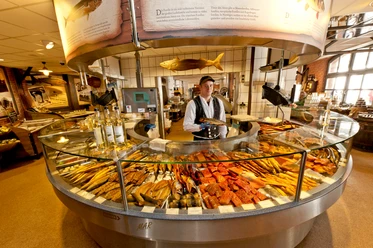KEY VISUAL 3-Fischmarkt Räucherecke_0092 (c) Fiedler_Erlebnis Bremerhaven.jpg Verkäufer hinter runder Theke mit großer Auswahl an frisch geräuchertem Fisch in Bremerhaven