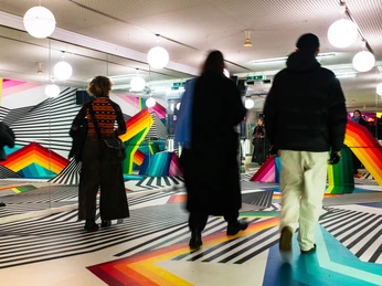 PASSAGEN - Interior Design Days Köln Lebendige Farben und geometrische Muster spiegeln sich in Spiegelwänden, während Besucher miteinander kommunizieren.Vibrant colors and geometric patterns are reflected in mirrored walls while visitors communicate with each other.