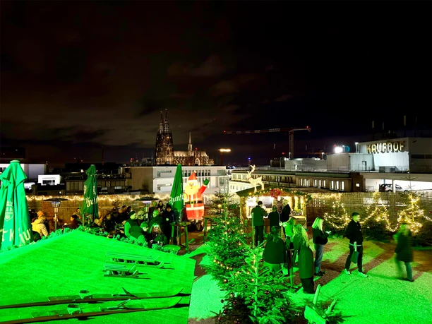 Nächtlicher Weihnachtsmarkt auf der Dachterrasse mit grünem Licht und Kölner Dom im Hintergrund.