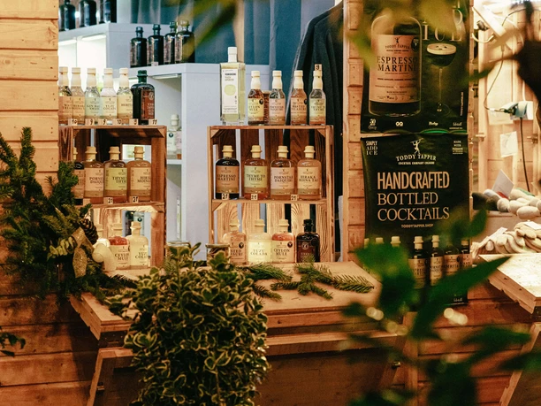 An illuminated wooden stand presents handmade cocktails and local products at a market.
