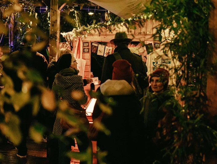 Winter market at the CBE Personen vor festlich geschmückten Ständen bei Nacht, umgeben von Lichtern und Dekor.People in front of festively decorated stalls at night, surrounded by lights and decorations.