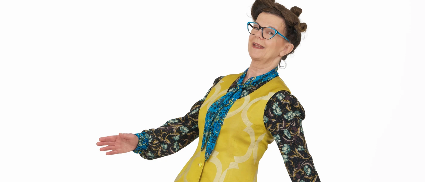 Frieda Braun_Auf ganzer Linie_hoch _Foto Britta Schüssling.jpg Frau mit blauer Brille in farbenfrohem Outfit lehnt sich lächelnd zur Seite vor weißem Hintergrund.