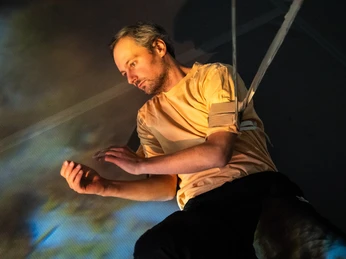 Ein Man, an Seilen befestigt, schwebt durch den Raum und klatscht in die Hände.A man, attached to ropes, floats through the room and claps his hands.