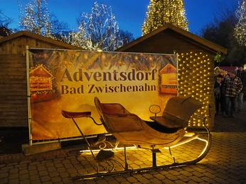 Markt-im-Advent-Adventsdorf-Schlitten.jpg Beleuchteter Schlitten vor Holzhäusern und Lichterbaum auf dem Adventsmarkt in Bad Zwischenahn