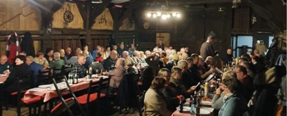 Moosetten.png Menschen sitzen in einem Holzsaal an langen Tischen beim gemeinsamen Grünkohlessen.