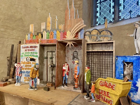 100 years of crib art at St. Gereon - Rudi Bannwarth building site crib Zu sehen ist eine moderne Interpretation der Weihnachtskrippe. Unter der Skyline von Köln sind diverse Personen aus der Stadtgesellschaft dargestellt: ein Fan des 1. FC Köln, ein Passant, eine Frau mit Kind, sowie eine Figur in der Arbeitskleidung eines Bauarbeiter und eine Frau mit Baby. Letztere sind offenbar die Interpretation von Maria und Josef.On display is a modern interpretation of the nativity scene. Various people from the city's society are depicted under the Cologne skyline: a fan of FC Köln, a passer-by, a woman with a child, a figure in the work clothes of a construction worker and a woman with a baby. The latter is apparently the interpretation of Mary and Joseph.