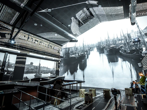 20230505 - Instawalk Fischereihafen 009 (c) Rainer Ganske_Erlebnis Bremerhaven.jpg Innenraum mit großflächiger Projektion historischer Fischkutter im Hafen, Besucher fotografieren die Szene.