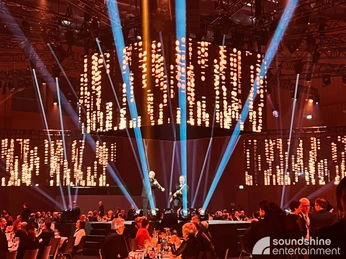 Show Produktion.jpg Bühnenshow mit zwei Musikern auf einer Würfelbühne, um die Gäste an Tischen herum sitzenStage show with two musicians on a cube stage, with guests seated at tables around them