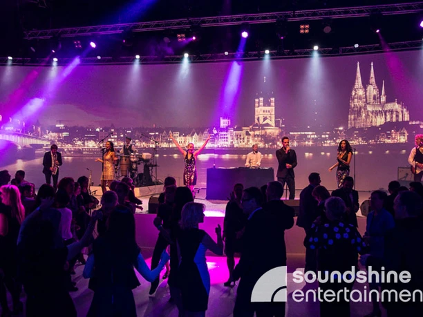 Soundshine Entertainment, Band, Cologne.jpg Eine Band steht auf einer Bühne mit einem Bühnenbild, das den Kölner Dom, die Altstadt und das Rheinufer zeigt
