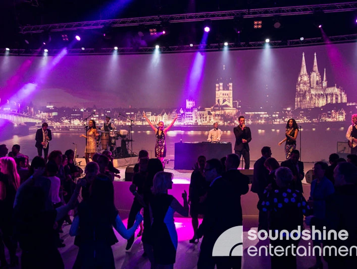 Soundshine Entertainment, Band, Cologne.jpg Eine Band steht auf einer Bühne mit einem Bühnenbild, das den Kölner Dom, die Altstadt und das Rheinufer zeigtA band stands on a stage with a stage set showing Cologne Cathedral, the old town and the banks of the Rhine