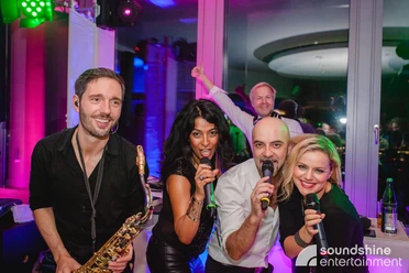 DJ Plus, Gesang, Saxofon.jpg 2 Sängerinnen, ein Sänger und ein Saxophonist lächeln in die Kamera