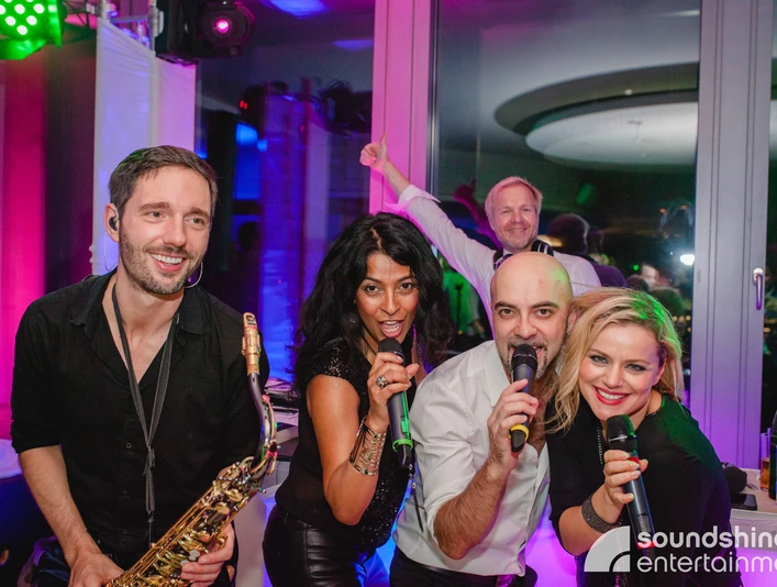 DJ Plus, Gesang, Saxofon.jpg 2 Sängerinnen, ein Sänger und ein Saxophonist lächeln in die Kamera2 female singers, a male singer and a saxophonist smile into the camera