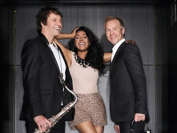 DJ Plus, Sängerin, Saxofon.jpg Ein Saxophonist und ein DJ in schwarzen Anzügen stehen neben einer schick gekleideten Frau mit schwarzen HaarenA saxophonist and a DJ in black suits stand next to a smartly dressed woman with black hair