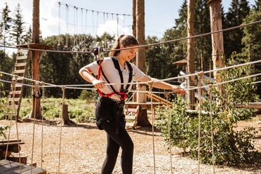 Kletterwald Surwold 1.jpg Person mit Klettergurt balanciert auf Seilen in einem Hochseilgarten zwischen Bäumen im Sonnenschein.