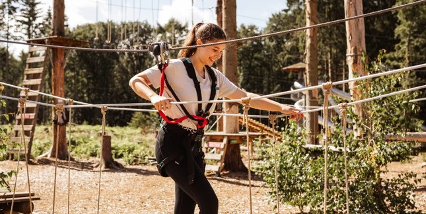 Kletterwald Surwold 1.jpg Person mit Klettergurt balanciert auf Seilen in einem Hochseilgarten zwischen Bäumen im Sonnenschein.