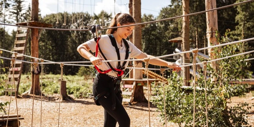 Kletterwald Surwold 1.jpg Person mit Klettergurt balanciert auf Seilen in einem Hochseilgarten zwischen Bäumen im Sonnenschein.