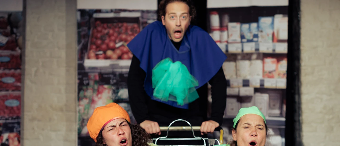 Horizont Theater Three people stand around a shopping cart with surprised expressions. They are wearing imaginative costumes.
