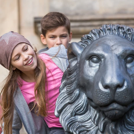 Zwei Kinder lehnen lachend an einer großen Löwenstatue in der Altstadt von Wolfenbüttel.