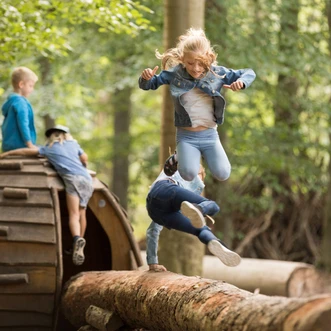 Kinder toben im Wald, springen von einem Holzstamm und spielen an einem großen Kletterelement.
