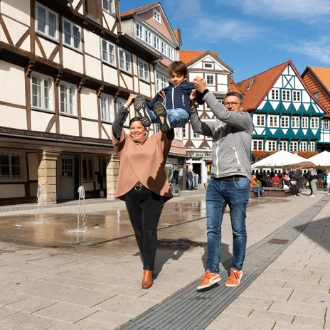 Familie hebt lachendes Kind auf Platz mit Fachwerkhäusern und Springbrunnen in Wolfenbüttel