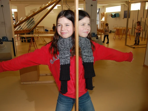 Phänomenta Spiegelungen (c) Phänomenta.JPG Ein Kind steht mit ausgebreiteten Armen vor einem Spiegel im Mitmachmuseum Phänomenta.