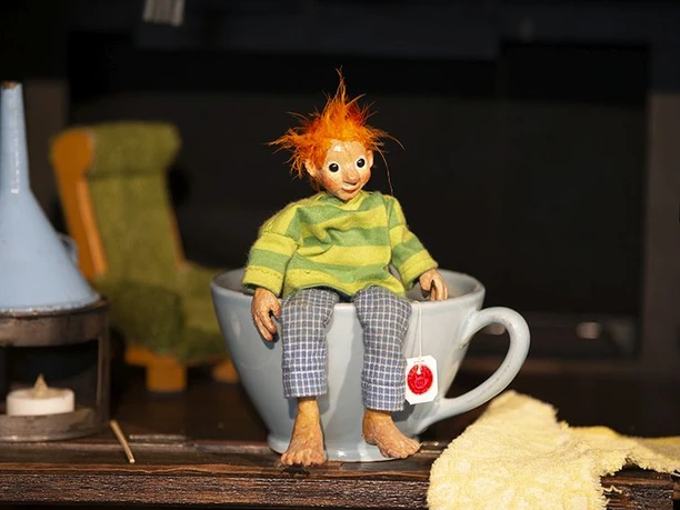 Holzfigur mit orangefarbenen Haaren und gestreiftem Pullover sitzt in einer Tasse auf der Bühne.