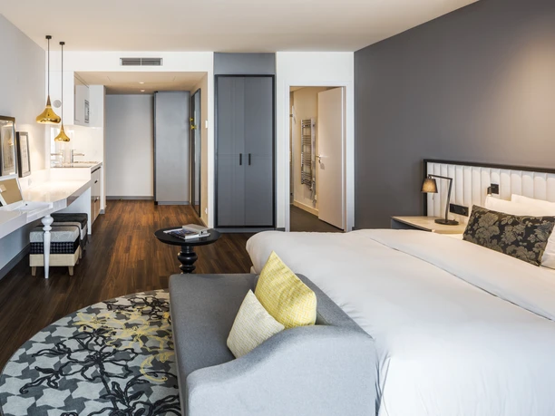 Capri by Fraser Frankfurt - Hotelzimmer Modernes Zimmer mit großem Bett, Schreibtisch und einem grauen Sessel am Fenster.