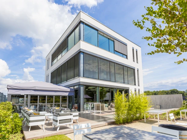 PERCUMA by CKE - Building view A modern, cubic white building with large glass fronts and external blinds.