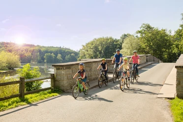 Familie mit Kindern radelt über eine Steinbrücke an einem bewaldeten Fluss.