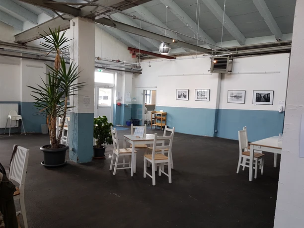 20201001_141349_Gretes Fischkai (c) Tanja Albert.jpg Innenraum eines Kulturcafés mit weißen Holztischen, Topfpflanzen und Fotografien an hellblauen Wänden.