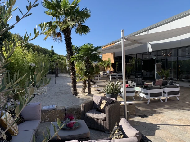 BOTANICAL by CKE - Außenterrasse Sonnige Außenterrasse mit sandigem Bereich, Palmen und bequemen Lounge-Möbeln, die eine entspannte Atmosphäre schafft.