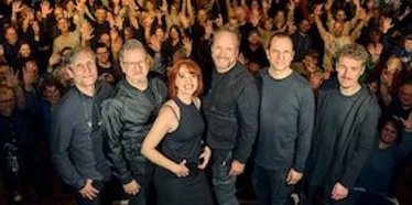 detailGroup_517680.jpg Sechs Musikerinnen und Musiker stehen auf einer Bühne vor einem jubelnden, dicht gedrängten Publikum.