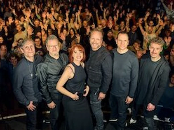 detailGroup_517680.jpg Sechs Musikerinnen und Musiker stehen auf einer Bühne vor einem jubelnden, dicht gedrängten Publikum.