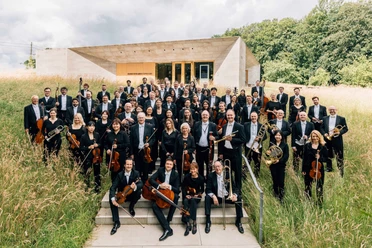 NWD Orchesterfoto Große Orchestergruppe in schwarzer Kleidung steht draußen vor modernem Gebäude im hohen Gras