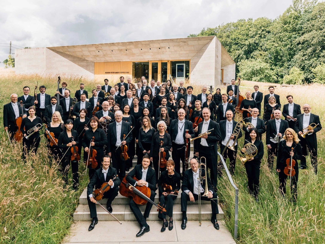 NWD Orchesterfoto Große Orchestergruppe in schwarzer Kleidung steht draußen vor modernem Gebäude im hohen Gras