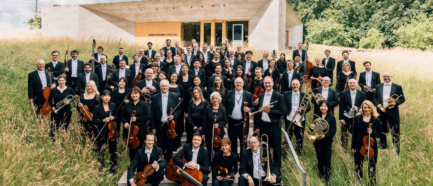 NWD Orchesterfoto Große Orchestergruppe in schwarzer Kleidung steht draußen vor modernem Gebäude im hohen Gras