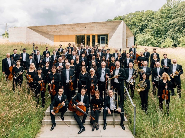 NWD Orchesterfoto Große Orchestergruppe in schwarzer Kleidung steht draußen vor modernem Gebäude im hohen Gras