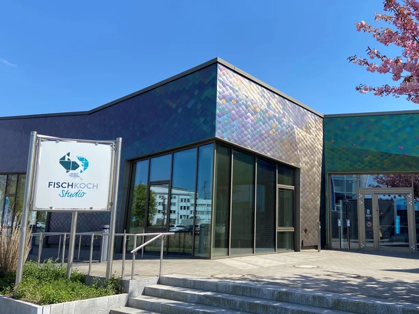 Fischkochstudio 2022_Außenansicht_FischKo (c) Christina Klug_Fischkochstudio.jpg Moderne Glasfassade des Fischkochstudios in Bremerhaven mit bunten Schuppenpanelen und Blütenbaum