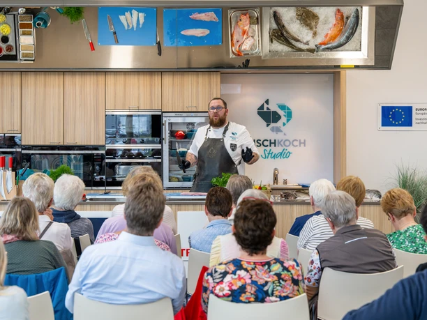© Björn Hake.jpg Koch erklärt vor Publikum die Zubereitung von Fischgerichten in moderner Studioküche in Bremerhaven.