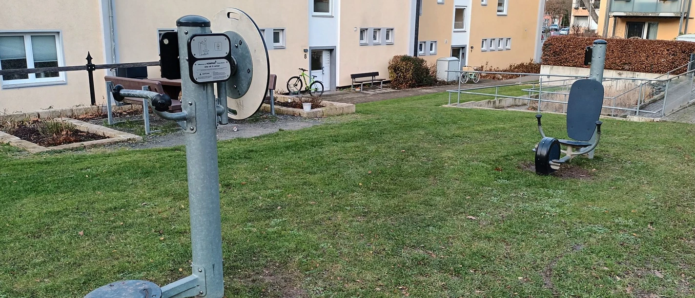 Fitnessgeräte_Eichendorffplatz_Salzgitter-Bad (7)_HD_16zu9.jpg Zwei Outdoor-Fitnessgeräte auf einer Rasenfläche vor mehrstöckigen Wohnhäusern in Salzgitter-Bad.