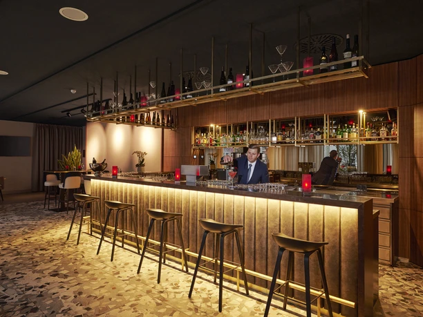 Frankfurt Airport - Hotel bar A modern hotel bar with an illuminated counter, high bar stools and a bartender in a suit preparing a drink. The ceiling is dark, the walls are wood-paneled.