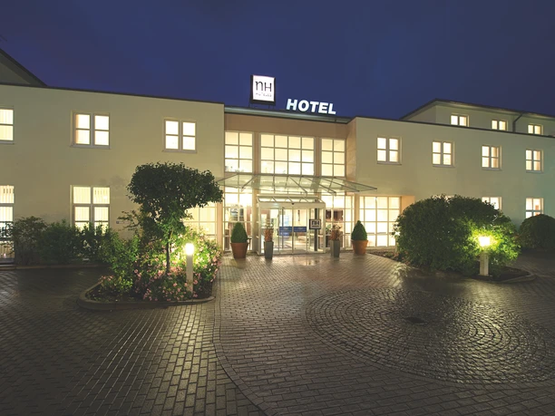 NH Frankfurt Airport - Hotel building An NH Hotel building at night, illuminated with warm light from the windows. The entrance area is surrounded by plants and lanterns.