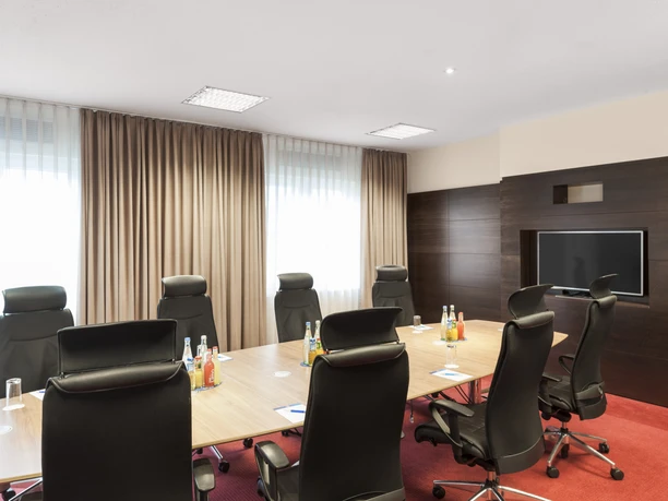 Frankfurt Airport - Konferenzraum Ein gut beleuchteter Konferenzraum mit einem Holztisch, schwarzen Stühlen und einem Fernseher an einer dunklen Holzwand.