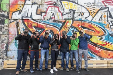 ctc events - Graffiti tour A group of seven people stand in front of a colorful graffiti wall. They are all holding a camera in front of their faces to photograph the wall.