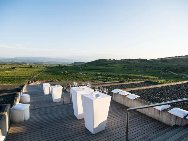 ctc events – Rheingau Terrace A terrace with white bar tables and wooden stools overlooks a vast hilly landscape with vineyards and a light blue sky.