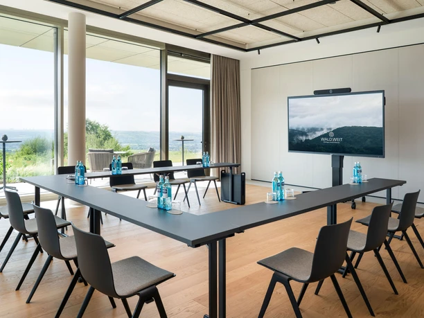 WALD.WEIT Rheingau Hotel & Retreat - WALD I Heller Tagungsraum mit bodentiefe Fenster, Parkettboden, U-Bestuhlung und Monitor.